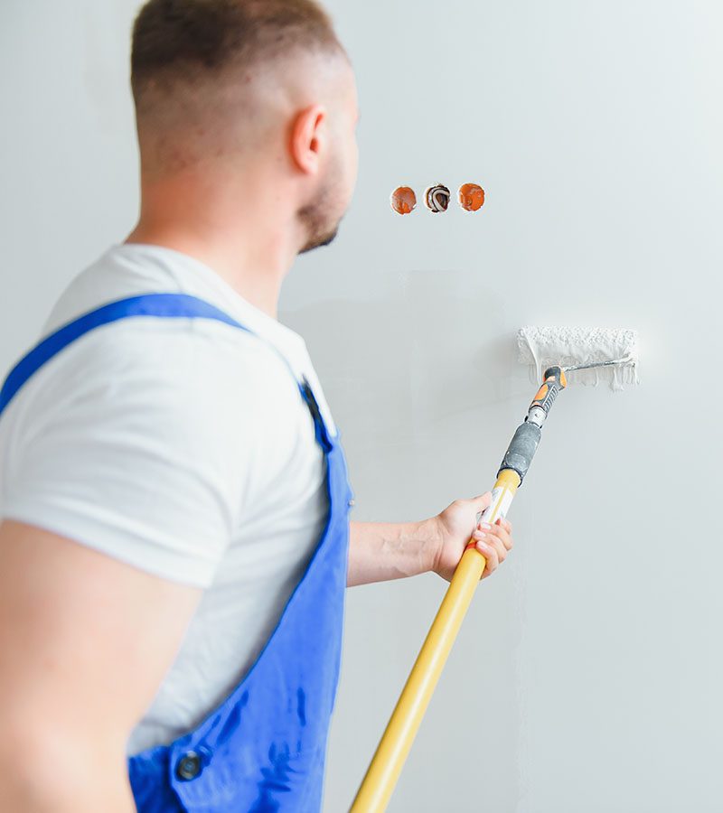 Workman in blue overalls painting the wall with a roller, view from the side